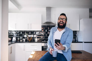 Freiberufler mit Smartphone und Kaffeetasse lachend zu Hause - ASGF02210