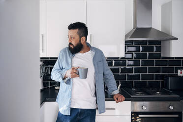 Nachdenklicher Mann mit Kaffeetasse in der Küche zu Hause stehend - ASGF02211