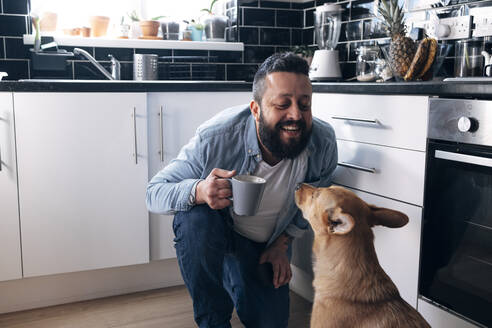 Glücklicher Mann mit Kaffeetasse, der einen Hund in der Küche betrachtet - ASGF02212