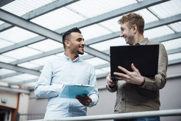 Smiling businessman holding laptop discussing with colleague at work place - JOSEF07817