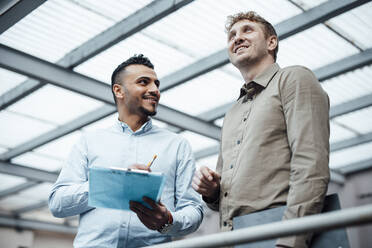 Happy businessman holding laptop standing with colleague at work place - JOSEF07818