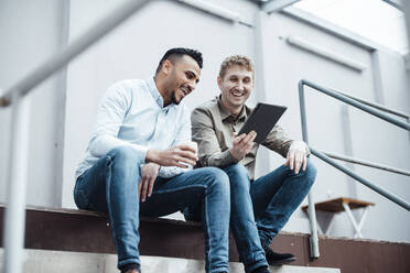 Happy businessman showing tablet PC to colleague siting by railing at work place - JOSEF07821