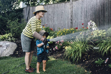 Happy grandmother with grandson watering plants together in garden - ACTF00175