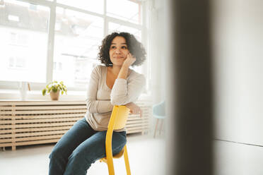 Smiling woman with hand on chin sitting on chair at home - JOSEF07835