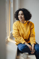 Smiling woman with coffee cup sitting by window at home - JOSEF07836