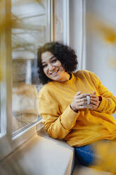 Smiling woman holding coffee cup leaning on window at home - JOSEF07837