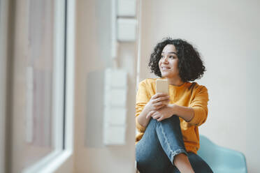 Thoughtful woman with mobile phone sitting by window at home - JOSEF07848