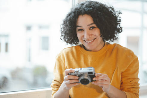 Lächelnde Frau mit Kamera, die zu Hause am Fenster sitzt - JOSEF07849