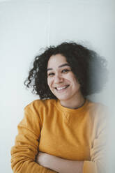 Happy woman with arms crossed standing in front of wall at home - JOSEF07852