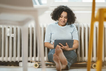 Smiling woman sitting on skateboard with tablet PC at home - JOSEF07855