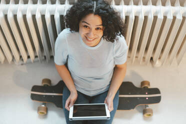 Glückliche Frau sitzt auf einem Skateboard mit Tablet-PC zu Hause - JOSEF07856