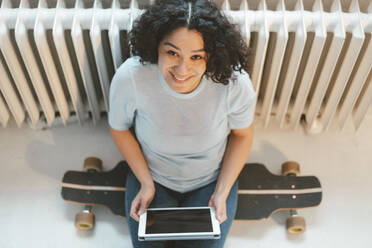 Happy woman sitting on skateboard with tablet PC at home - JOSEF07856