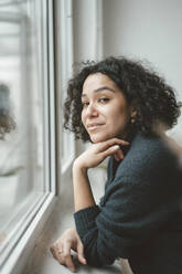 Beautiful woman with curly hair standing by window at home - JOSEF07864