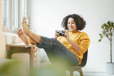 Smiling woman with camera sitting on chair at home - JOSEF07872