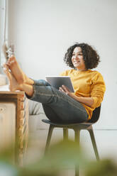 Happy beautiful woman sitting with tablet PC on chair at home - JOSEF07874