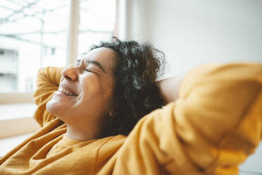 Cheerful woman with hands behind head sitting at home - JOSEF07887