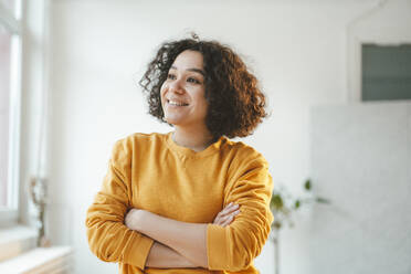 Smiling thoughtful woman standing with arms crossed at home - JOSEF07892