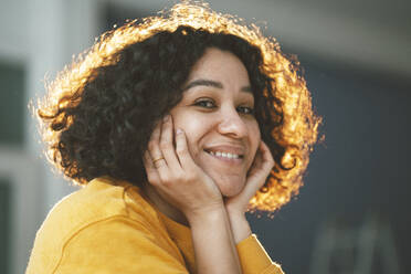 Happy beautiful woman with curly hair sitting with head in hands at home - JOSEF07897