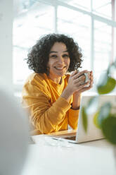 Happy woman holding coffee cup sitting with laptop at table - JOSEF07900