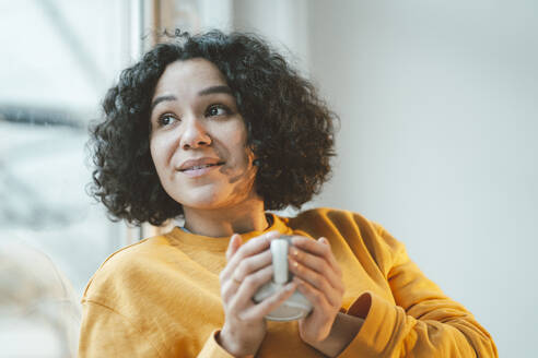 Nachdenkliche Frau mit lockigem Haar hält Kaffeetasse zu Hause - JOSEF07903