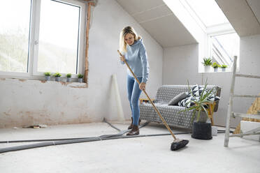 Smiling blond woman sweeping floor with broom in attic - HMEF01344