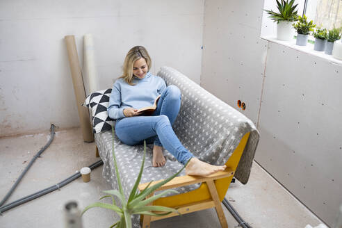 Lächelnde blonde Frau sitzt auf dem Sofa und liest ein Buch auf dem Dachboden - HMEF01346