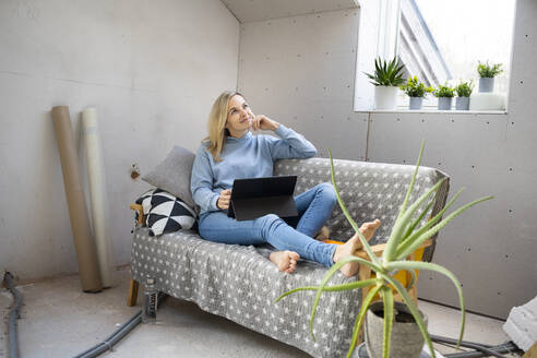 Nachdenkliche blonde Frau mit Tablet-PC auf dem Sofa in der Mansarde sitzend - HMEF01352