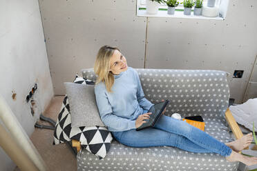 Glückliche blonde Frau mit Tablet-PC auf dem Sofa in der Mansarde sitzend - HMEF01353