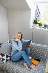 Happy woman with hand on chin sitting on sofa with tablet PC in attic - HMEF01354
