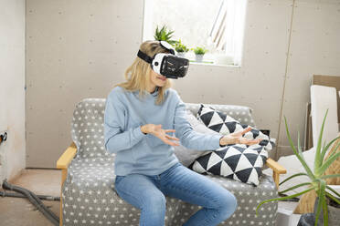 Smiling blond woman gesturing with virtual reality simulator sitting on sofa in attic - HMEF01357