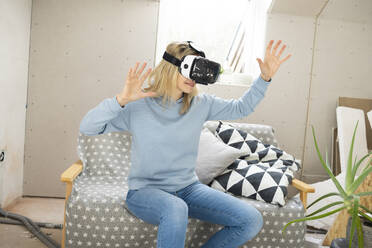 Lächelnde Frau gestikuliert mit einem Virtual-Reality-Simulator auf einem Sofa im Dachgeschoss - HMEF01358