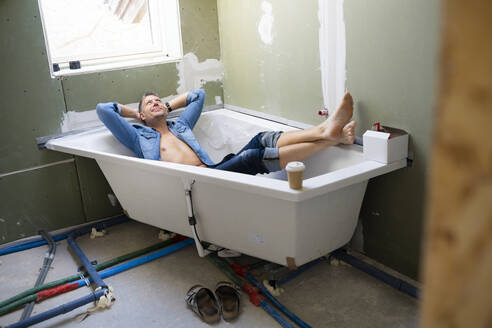 Lächelnder Mann mit Händen hinter dem Kopf in der Badewanne auf dem Dachboden liegend - HMEF01359