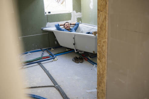 Happy man with hands behind head lying in bathtub in attic - HMEF01360