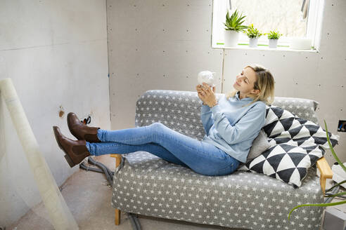 Lächelnde blonde Frau mit Sparschwein auf dem Sofa in der Mansarde sitzend - HMEF01363