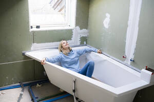 Glückliche blonde Frau sitzt in der Badewanne auf dem Dachboden - HMEF01370