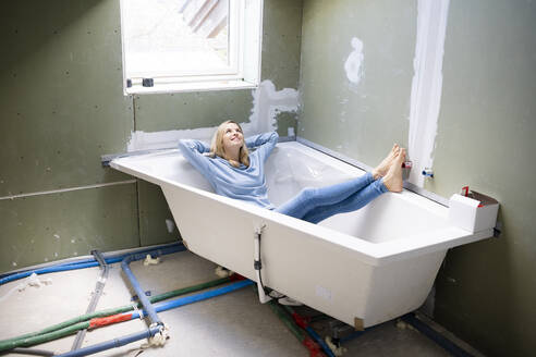 Happy blond woman with hands behind head sitting in bathtub in attic - HMEF01371