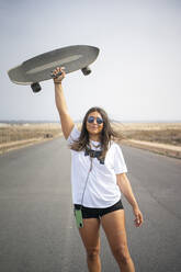 Lächelnde Frau mit Sonnenbrille hält Skateboard auf der Straße hoch - FBAF01866