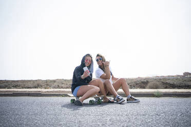 Friends taking selfie through smart phone by sitting on skateboard on sunny day - FBAF01873