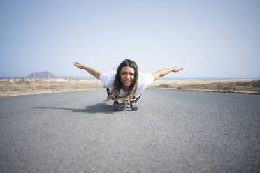 Frau mit ausgestreckten Armen auf dem Skateboard liegend - FBAF01878