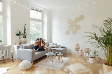 Woman listening music through headphones sitting with daughter on sofa at home - JOSEF07907