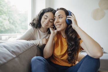 Glückliche Frau hört Musik über Kopfhörer und sitzt mit ihrer Tochter auf dem Sofa zu Hause - JOSEF07910