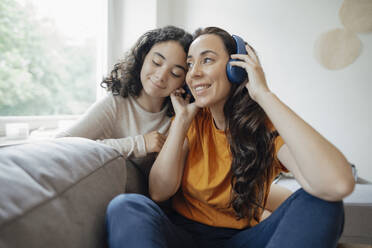 Happy woman listening music through headphones sitting with daughter on sofa at home - JOSEF07910