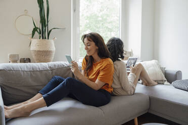 Smiling woman using smart phone sitting back to back with daughter using tablet PC on sofa at home - JOSEF07951