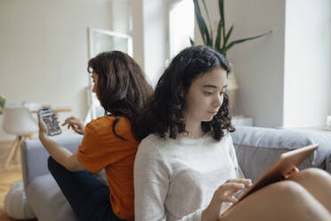 Teenage girl using tablet PC sitting back to back with mother holding mobile phone - JOSEF07953