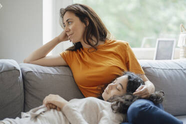 Teenage girl resting in lap of mother on sofa at home - JOSEF07955