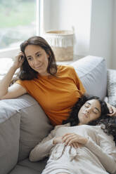 Teenage girl resting in lap of thoughtful mother on sofa at home - JOSEF07956