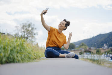 Smiling woman gesturing pace sign taking selfie on wall - JOSEF07961
