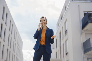 Smiling businesswoman talking on mobile phone in front of buildings on sunny day - JOSEF07965