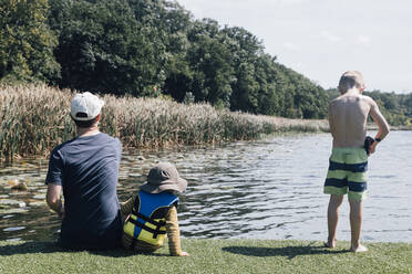 Vater mit Söhnen am Flussufer sitzend an einem sonnigen Tag - ACTF00181