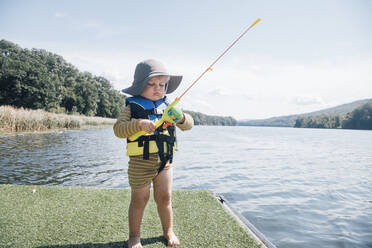 Cute baby boy with fishing rod standing by river on sunny day stock photo